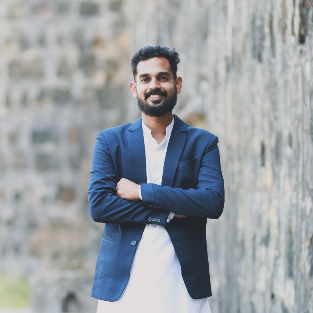 Young south asian man smiling confidently in a blue suit against a stone wall background.