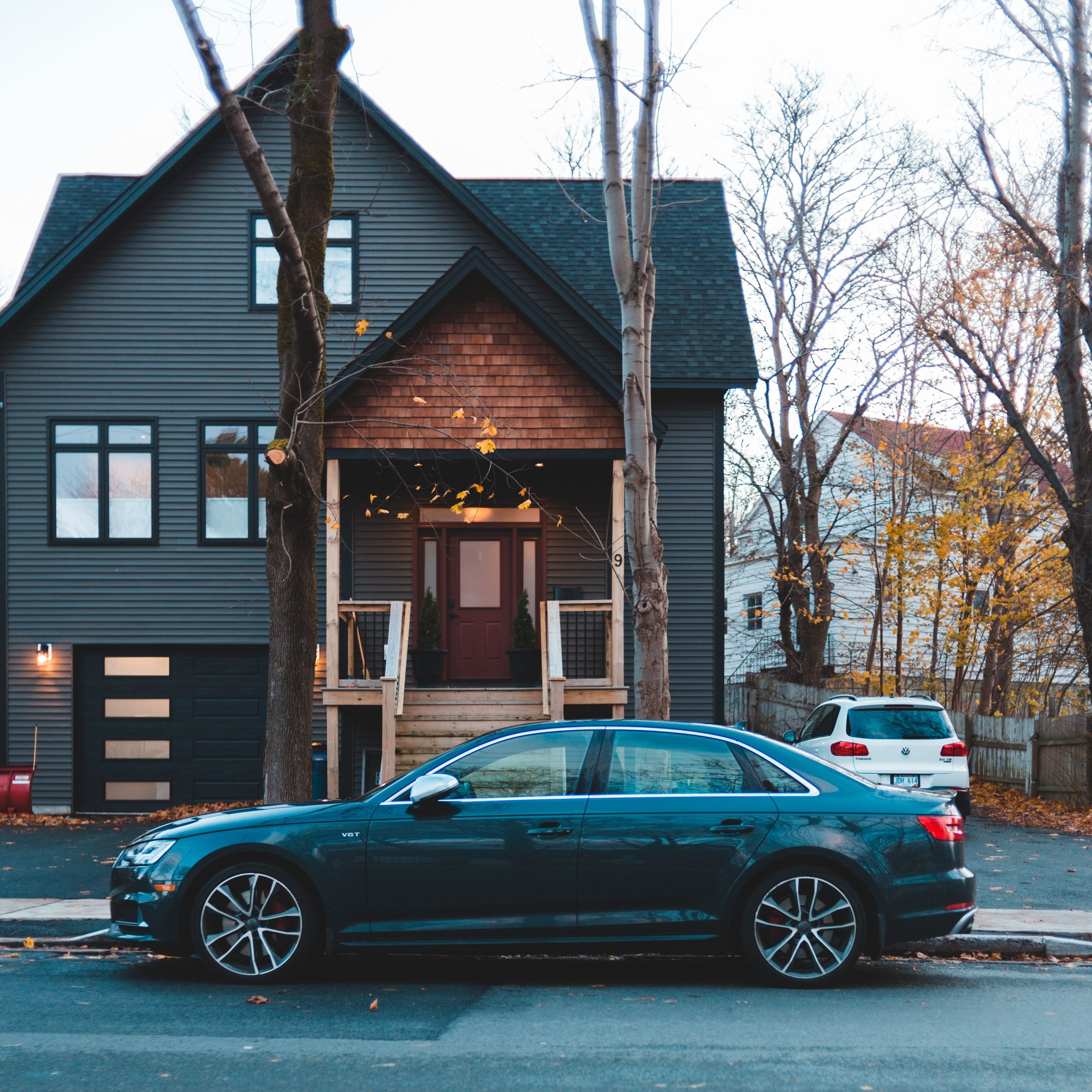 A classic car parked on a street in front of a modern dark-colored house, in an autumn setting.